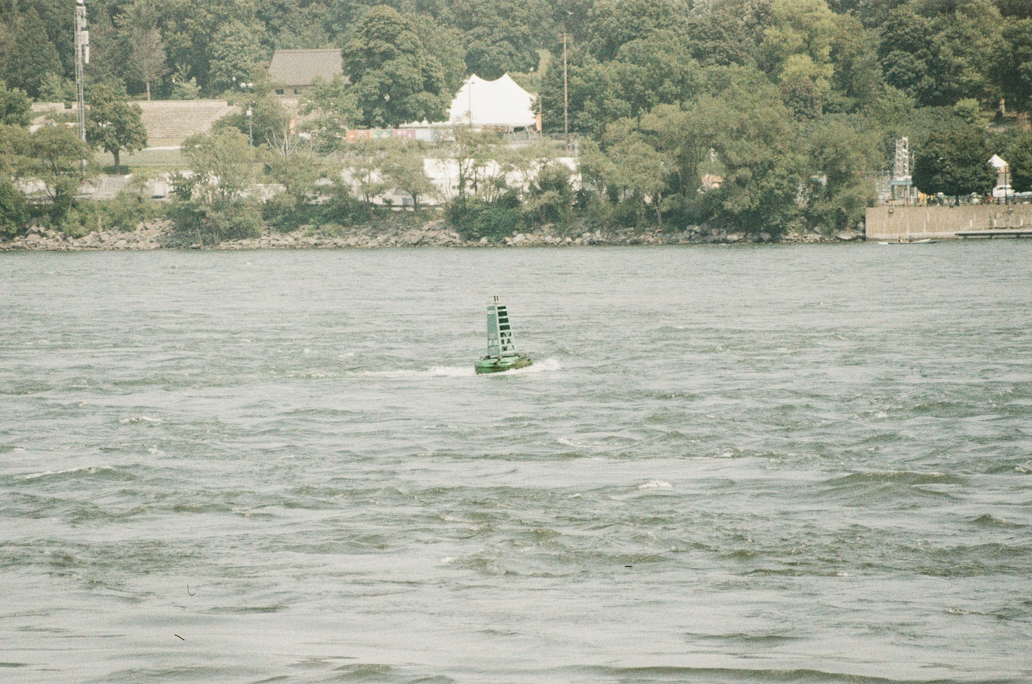 a green buoy in the middle of the river.
the far shore is rocky with some trees.