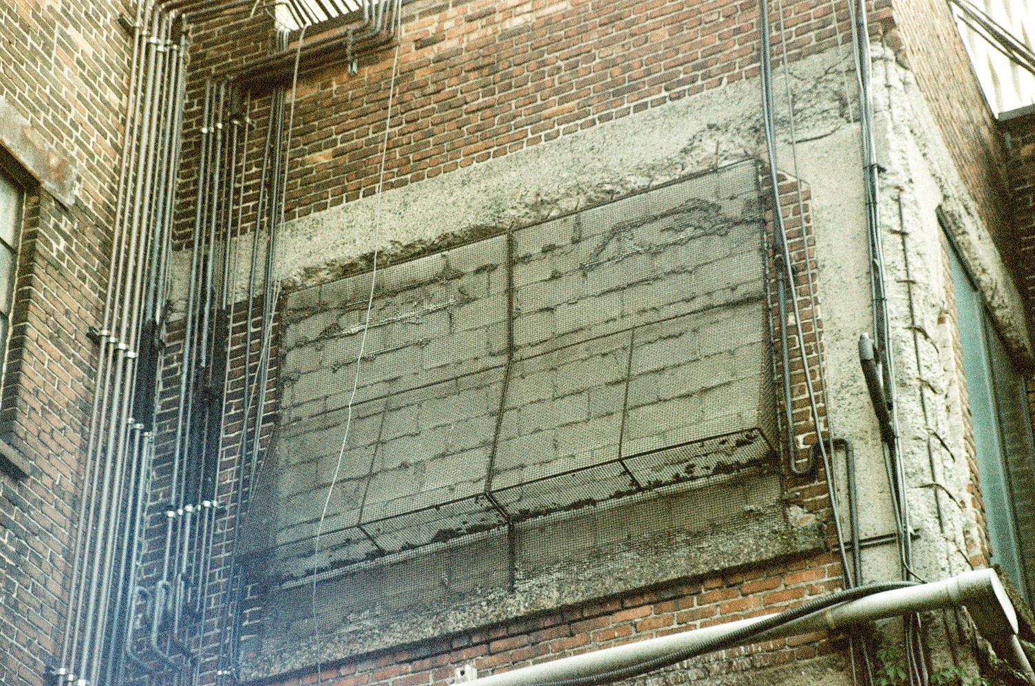 part of an old factory building,
a storey or two up.
what used to be a window
has been filled in with big grey stone bricks,
but the metal cage over the outside
has been left in place.
a whole array of pipes climbs
the building to the left.