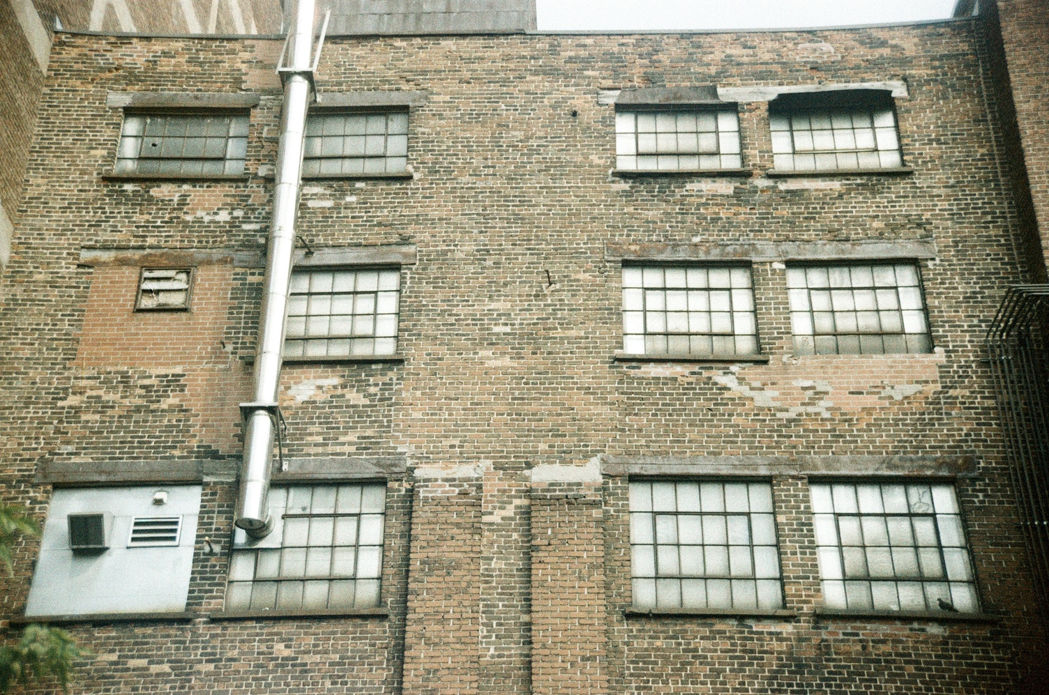 the brick wall of an old factory building
with sets of 6x4 pane windows,
where it looks like the middle 4x2
panes swivel open.
a round duct or chimney
has been installed
in one of the lower windows
and goes up the outside of the building.
two of the other windows
have been filled in,
one with brick
and one with metal.
they both have vents embedded in them.
an array of pipes climbs halfway up
the far right edge of this wall.