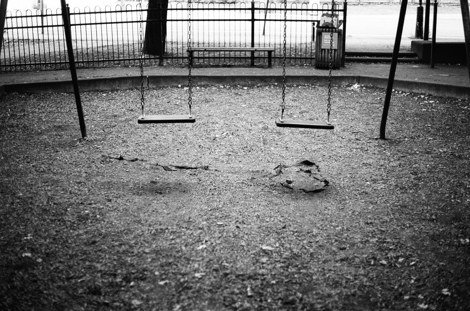 two empty swings in a park.
beyond there is a bench and a trash can.