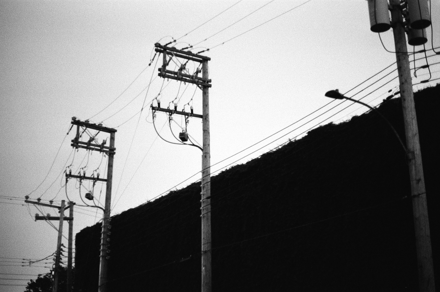 a series of 4 power line poles
in close proximity
against a grey sky
in front of a completely shadowed building.