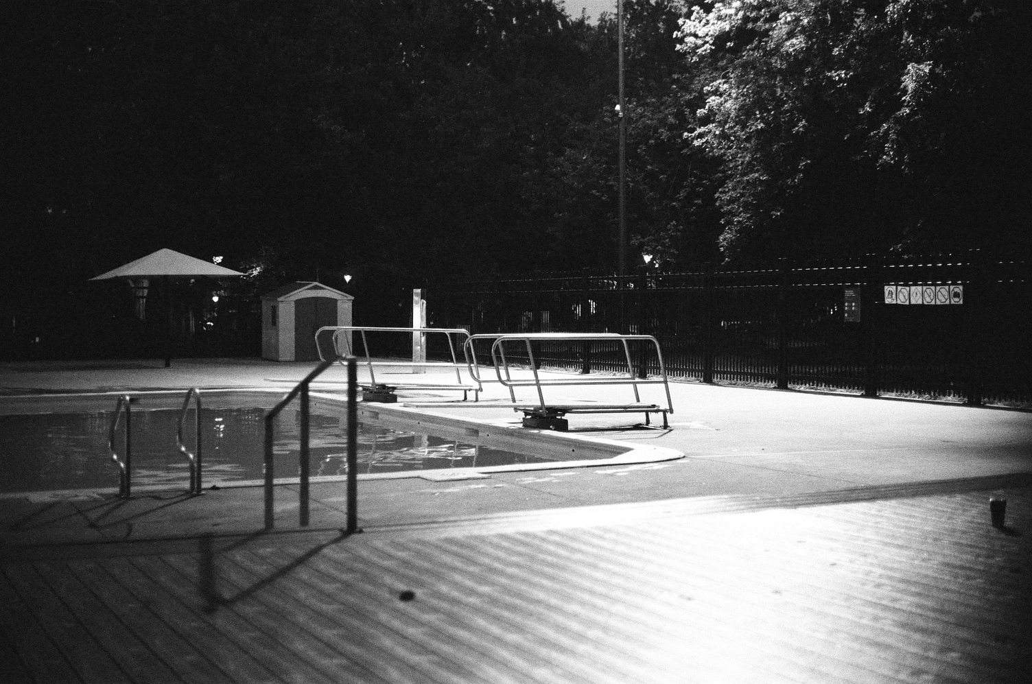 a public pool in a park
at night illuminated by flood light above.
in the centre of the frame
are two diving boards side by side
with metal railings.