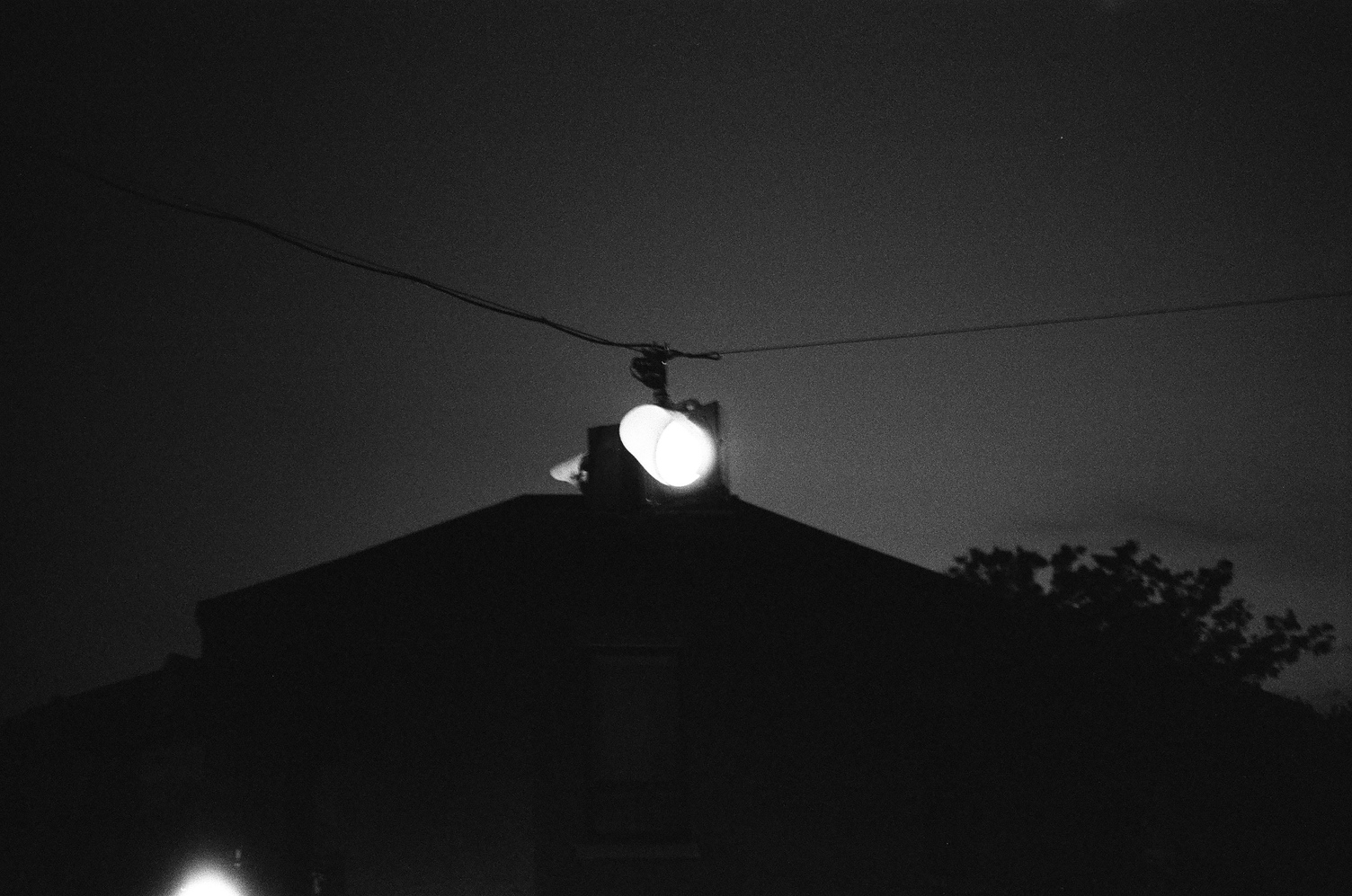 a blinking red traffic light suspended above an intersection.
it's completely dark
except for the traffic light
and some other source in the bottom left.
the shape of a building and tree can be made out.