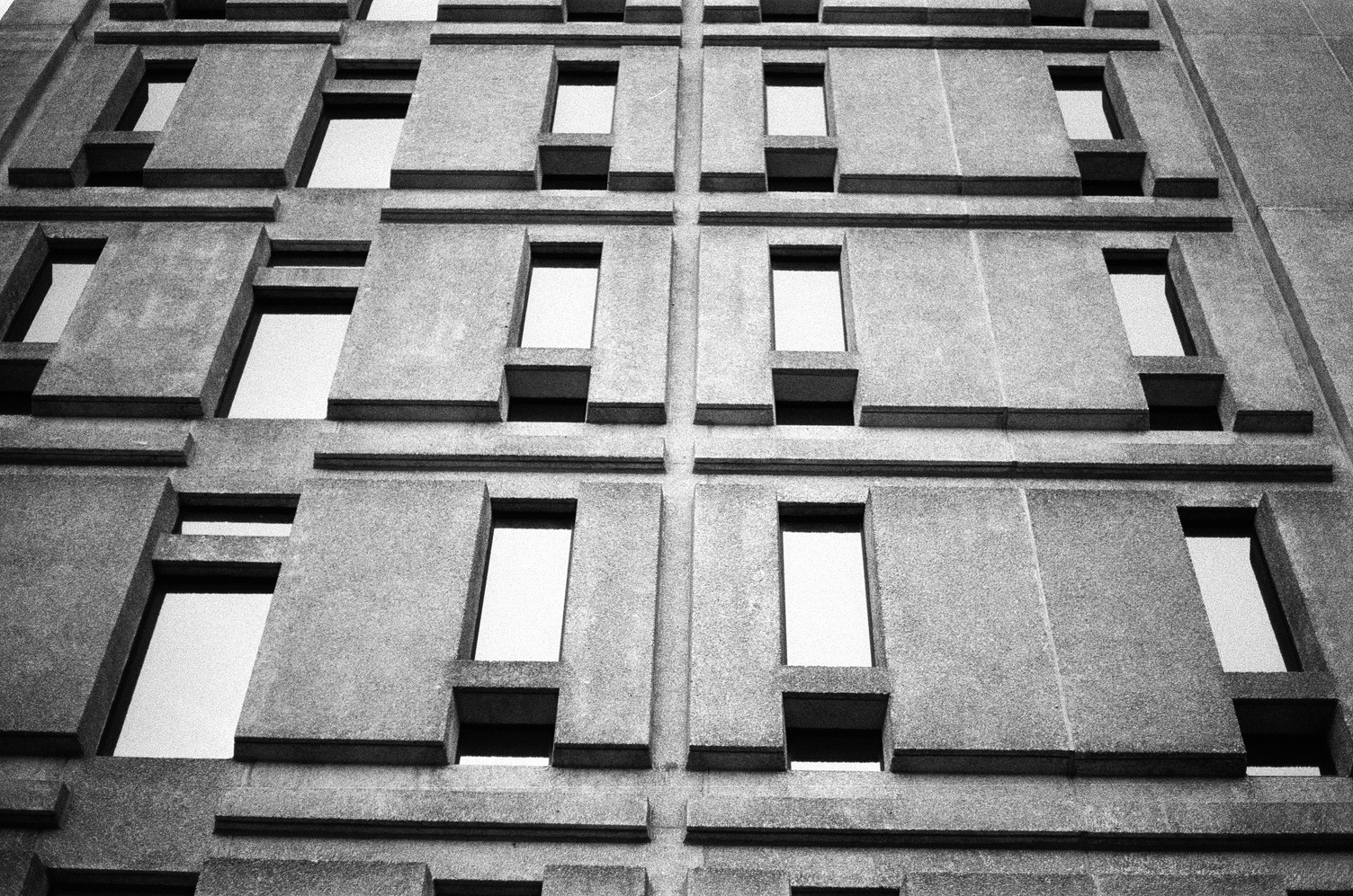 a wider view of
the side of a concrete building
with an intricate pattern
of concrete and narrow windows.
the windows are reflecting a light sky.
good contrast and shadows here.