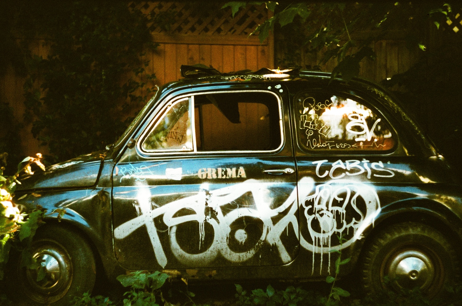 the side of a weird old presumably european tiny car
that's been rotting in an alley for who knows how long.
the car is painted black
and there's a bunch of graffiti in white
on the door and back side as well as the window.
on the door next to the handle there's a stenciled "GREMA".
the car is in worse shape
than the last time I photographed it.