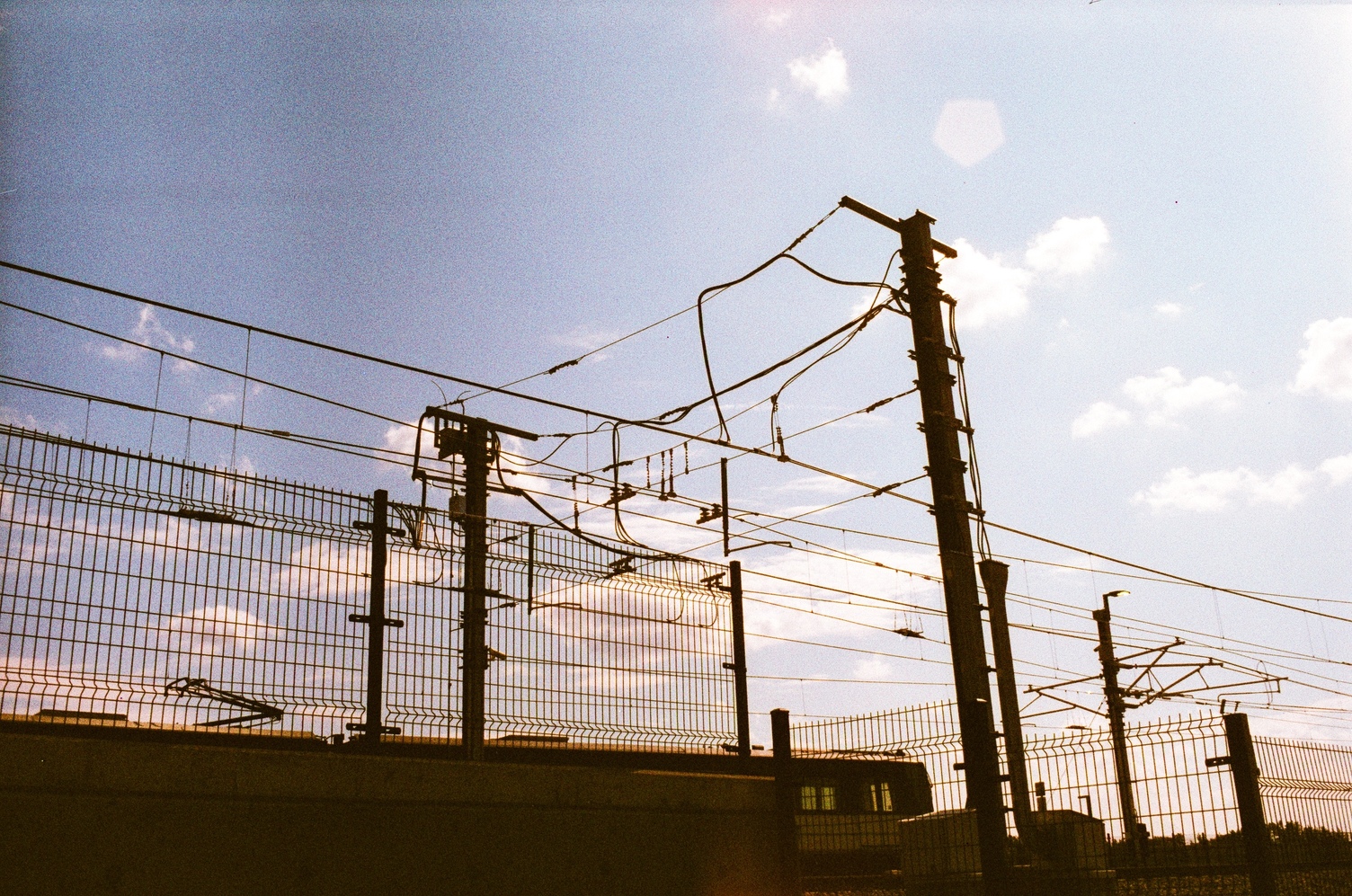 overhead lines for electric trains
silhouetted against a blue sky.
there's a train partially visible
behind a concrete barrier.
there's a pentagonal lens flare
in the sky.