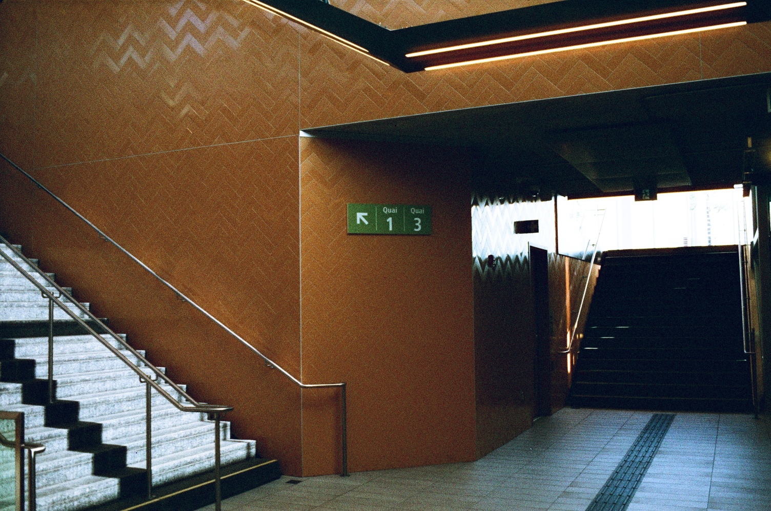 the inside of a train station,
with walls of orange-brown tile.
there is a stairway leading up to the left,
and one in shadow at the end of a short tunnel
to the right.
in the centre is a green sign
pointing up the left stairs towards
quai 1 and quai 3.