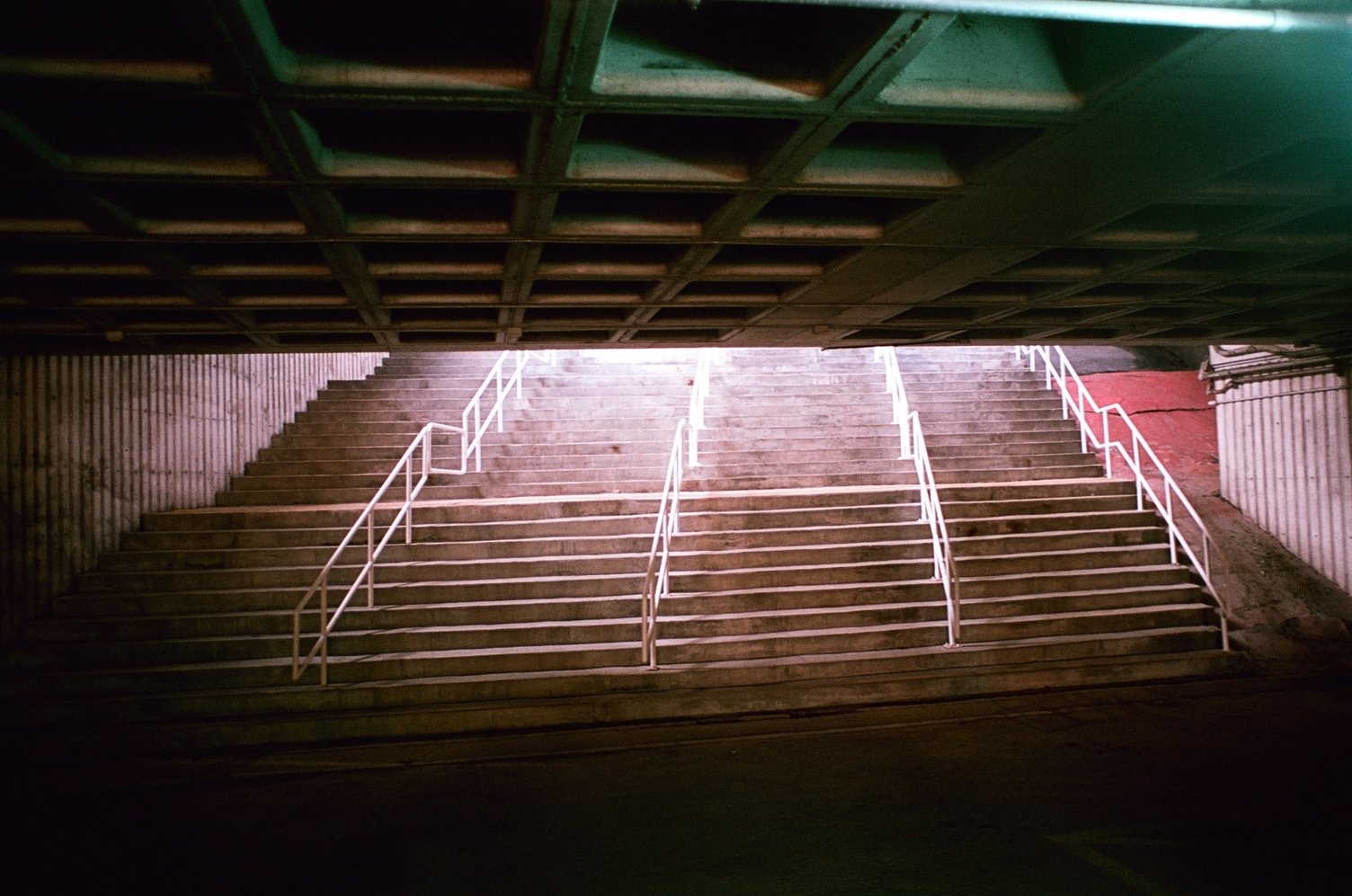 a wide set of concrete stairs
divided by 4 sets of white metal railings
ascending out into the sun
from under a low concrete ceiling
with a large square grid pattern.
the ceiling is being lit
by a sort of bluish-green light
from behind.