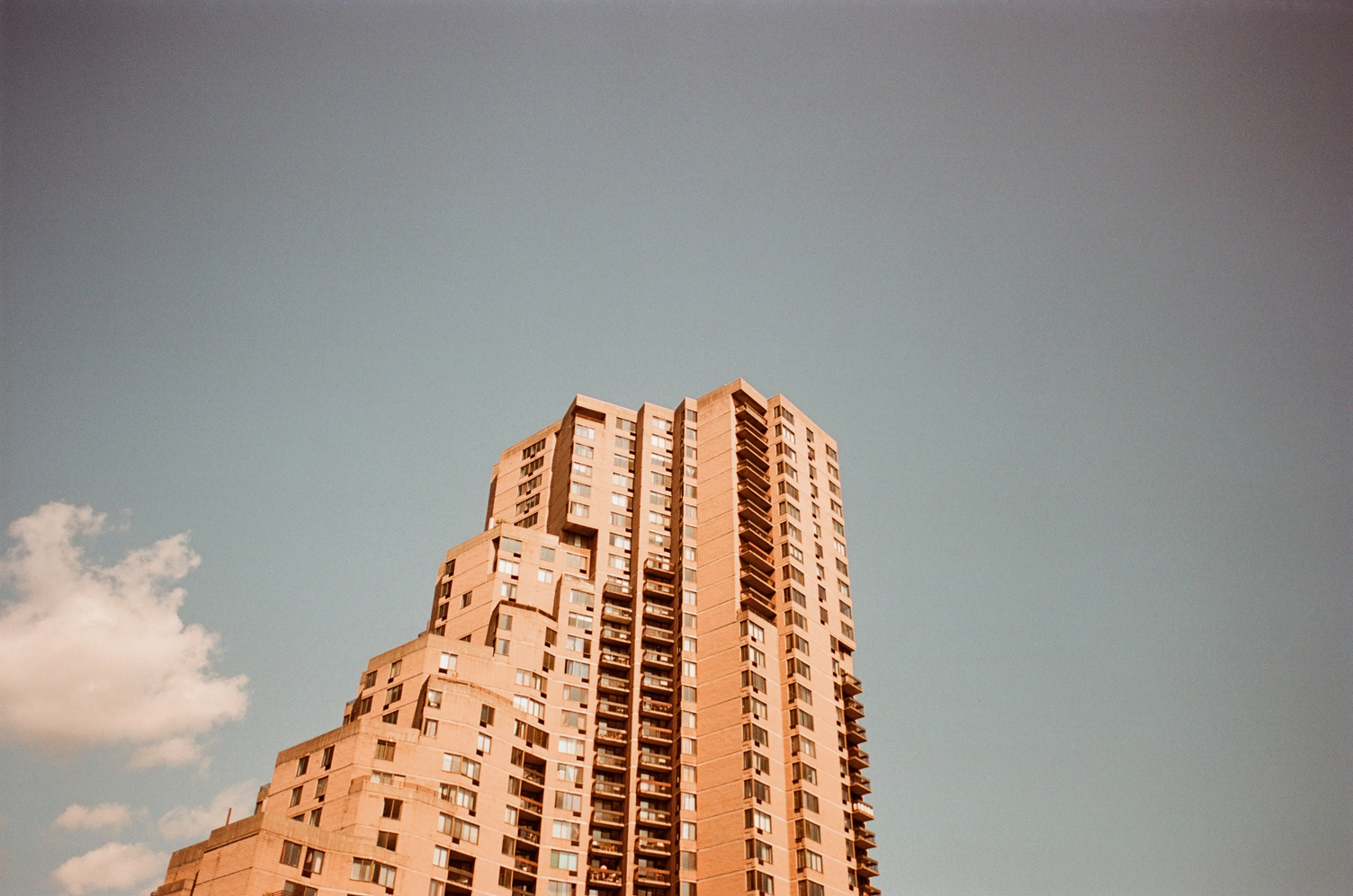 a tall brutalist-ish apartment building
on a mostly clear blue sky,
with one white cloud in the bottom left.
the building appears orange.