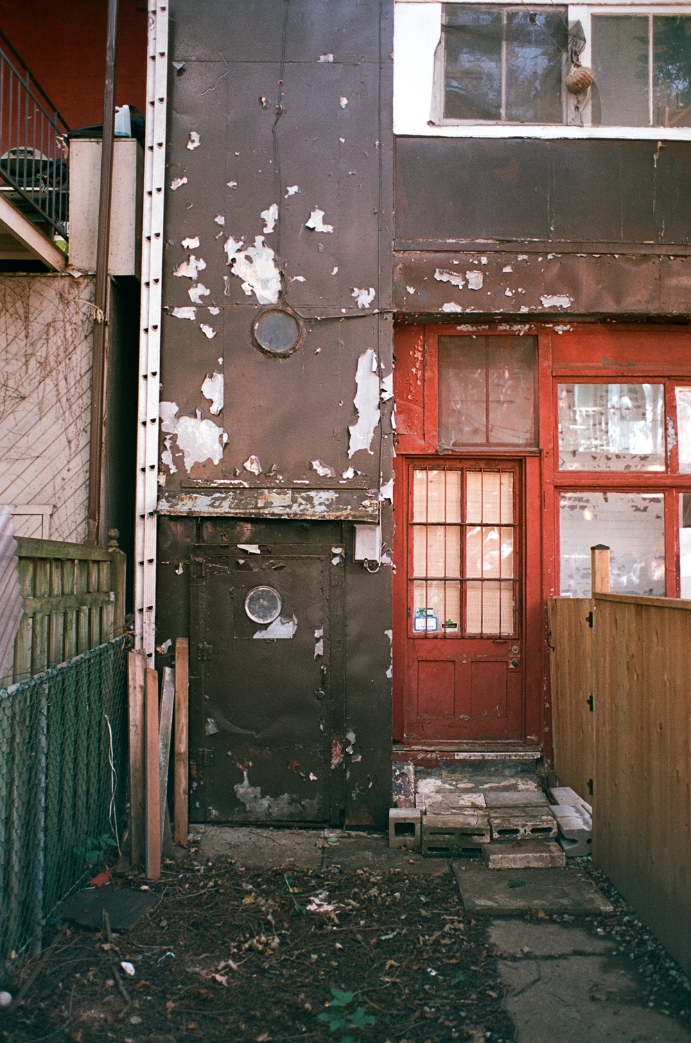 the back of a very strange
residential building
that's clearly been built onto.
on the right is a regular red wooden door.
on the left is a painted brown metal door
with a circular window in it
like a porthole.
this is at the bottom of a metal column,
presumably containing stairs,
with one more porthole
not far above the door.
slid right between this weird extension
and the side of the adjacent building
is a very tall ladder.