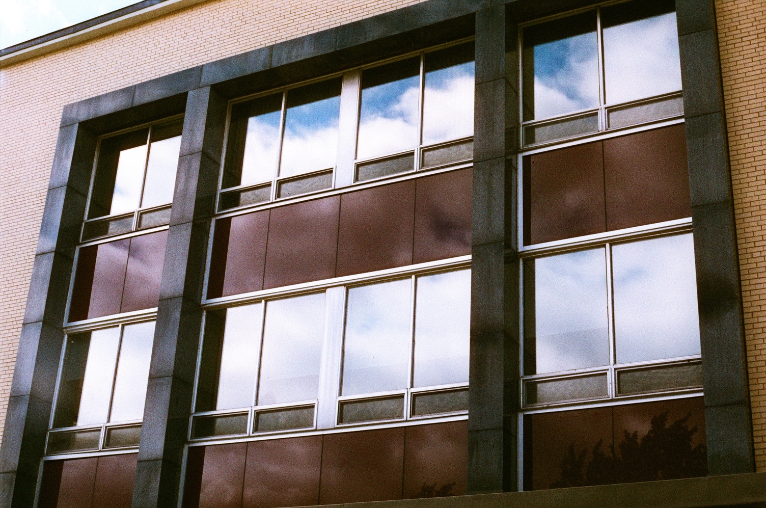 a block of windows in a school building,
viewed from a slight angle.
the wall of the building is beige brick
and the block of windows
is bordered by grey stone.
there are two rows of windows,
and below each row are panels that appear brown,
though they're orange in person.
the top row of windows
is reflecting some blue sky and clouds,
while the bottom row is reflecting only cloud.