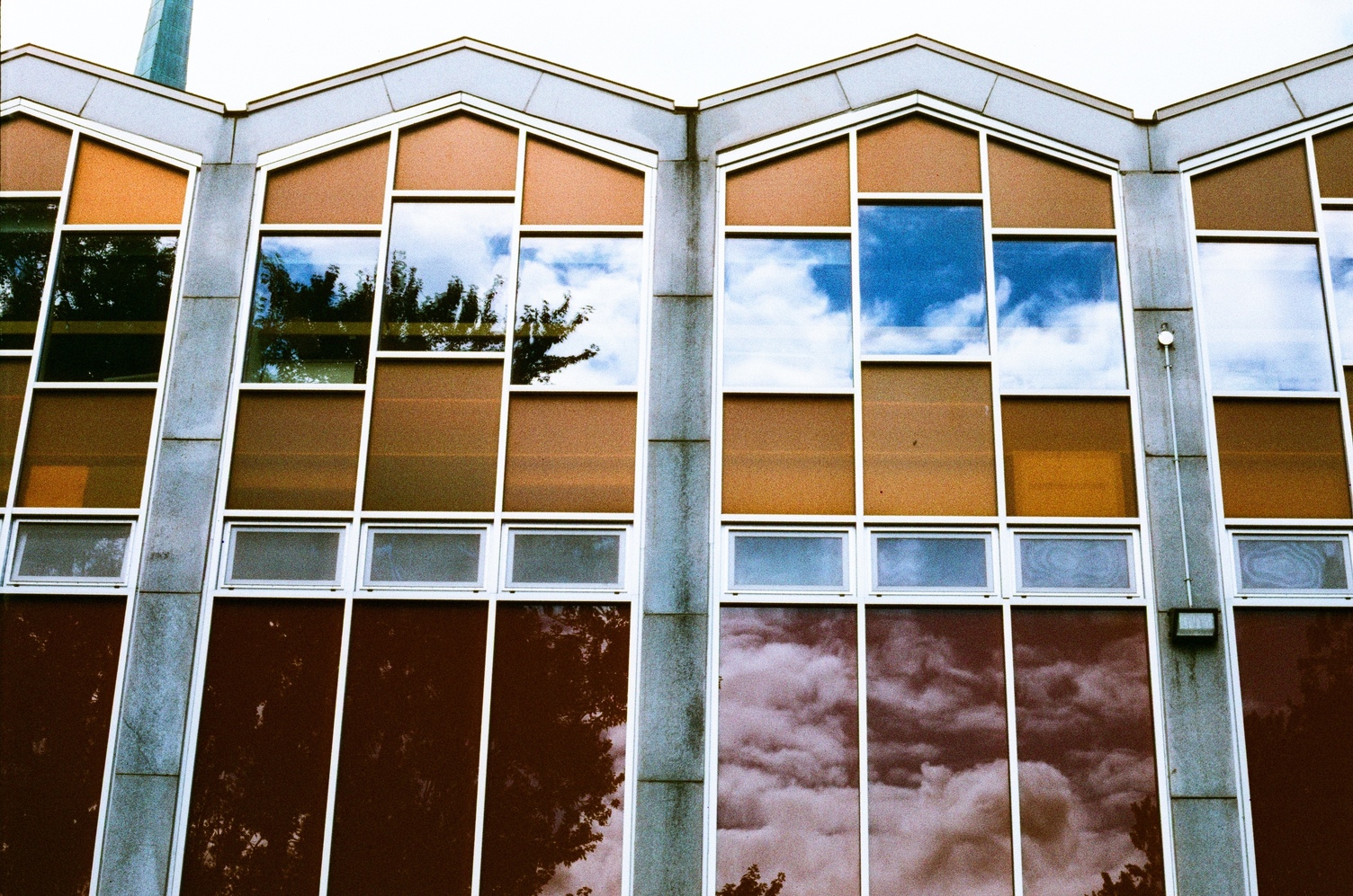 fancy windows on the side of a school building.
it's a repeating pattern of three windows
in the middle of a house-shaped design
of yellow panels on the second floor.
the middle windows are higher than the other two.
the triangle at the top of the window design
is followed by the profile of the roof.
below that, separated by a row of even short windows,
are red panels.
the upper windows are reflecting blue sky and clouds,
while the lower red panels are reflecting trees and clouds.