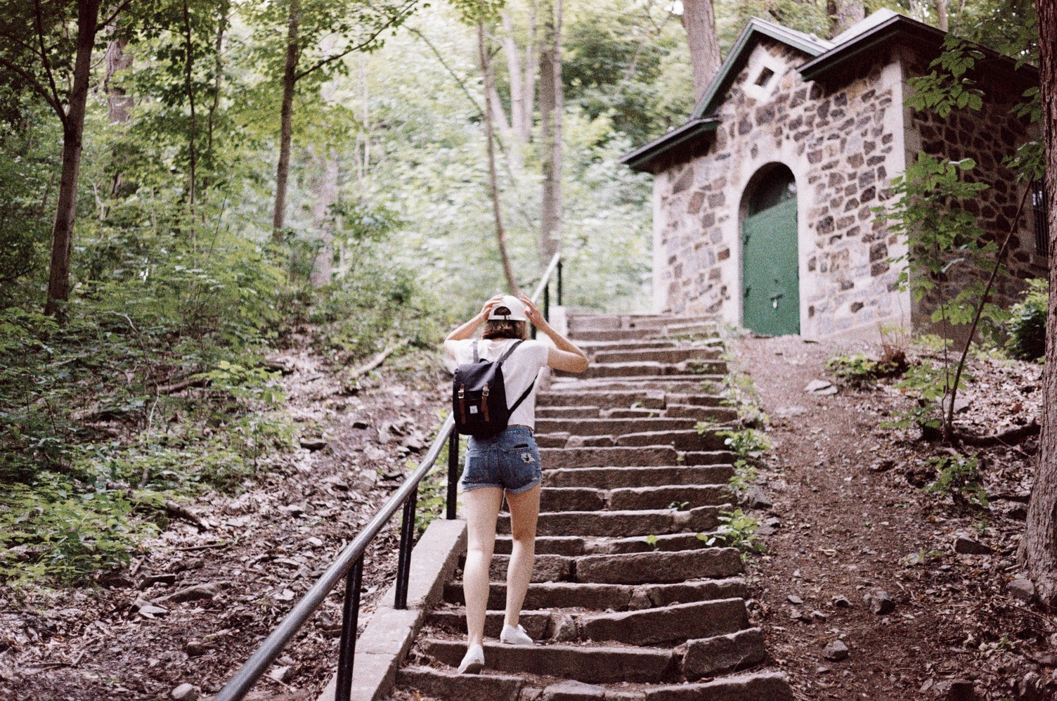 Ayla in a light coloured tshirt,
short jorts,
small backpack,
white baseball cap
from behind walking up stone steps
towards a small stone building
with a green door.