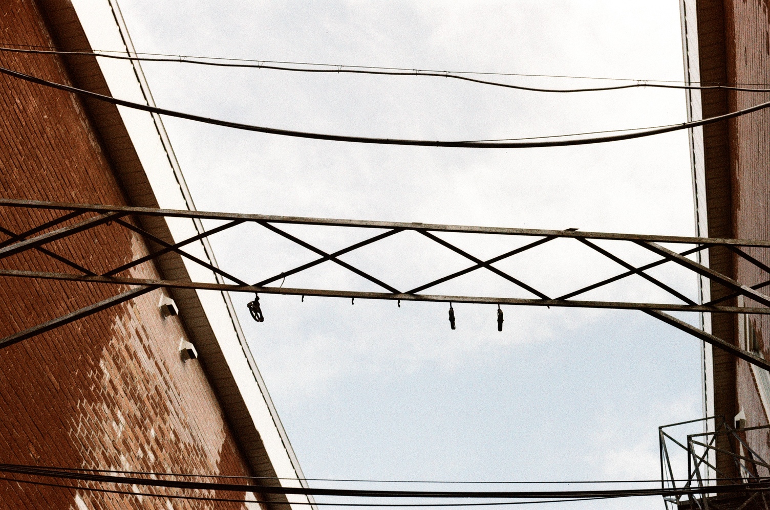 a view up between two buildings
at a metal truss in silhouette
against the almost white sky
and some cables running between them.