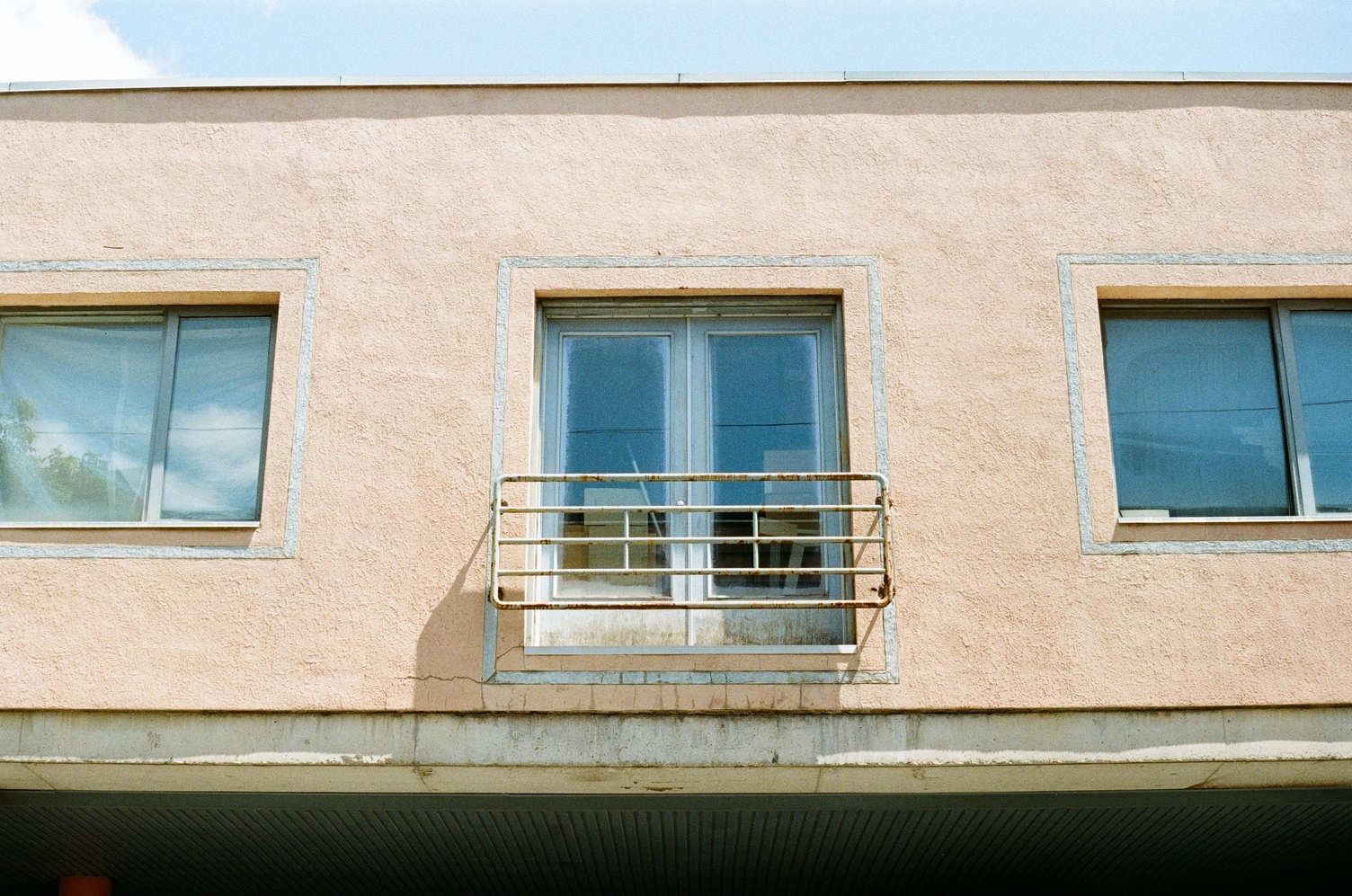 a floor of a building raised on stilts,
though they aren't in frame.
it's a beige plaster wall
with a set of doors
in the centre
guarded by just a fence
with no balcony.
the doors are flanked
by windows on either side.
the doors and windows
have oversized outlines in grey.