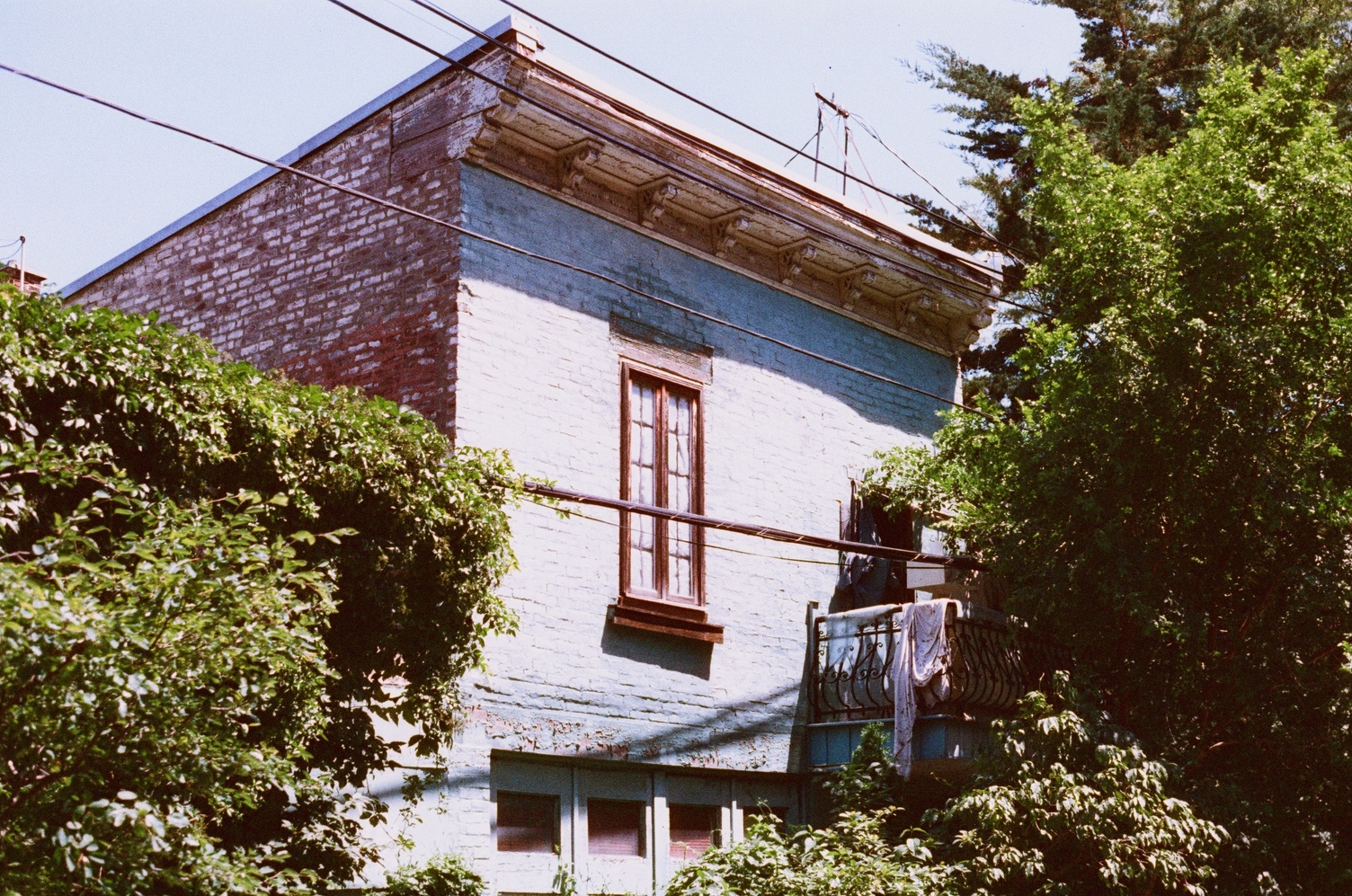 a brick house crowded by trees.
the front face is painted a light blue.
there's a tall narrow window
above a garage door,
and a balcony
with some cloth draped over its railing.