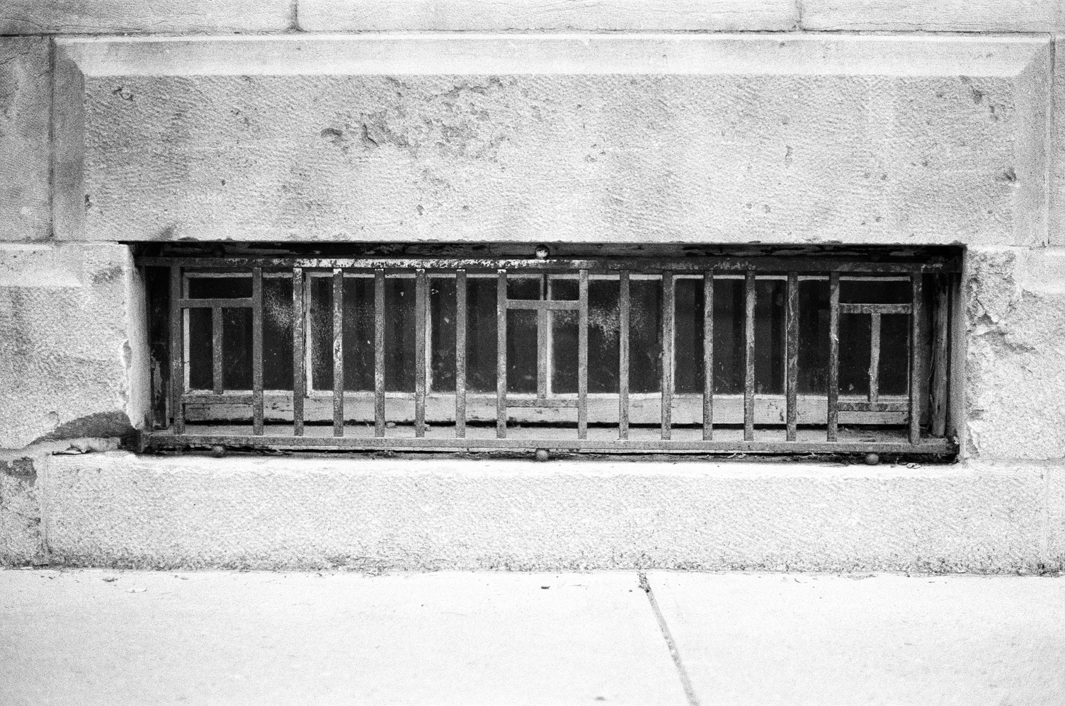 a low,
short and wide
window into a basement
in a stone wall.
there is a metal grate
in front of the window
with a pattern of three
sideways H shapes
separated by straight vertical bars.