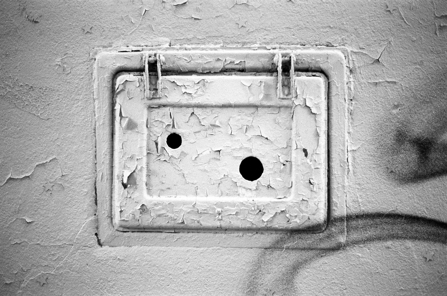 a small horizontal metal door
in a plaster wall
with very flaky paint.
there are two holes
in the small door,
a smaller one in the top left
and a larger one in the bottom right.
there are stars,
possibly stickers,
placed around the opening.