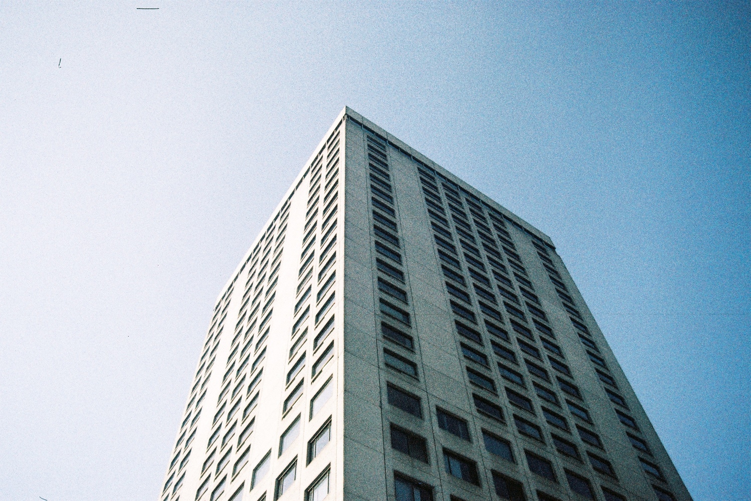 looking up at the corner of a grey office building
of at least 18 floors that we can see,
on a clear blue sky.
the sun is on the left,
illuminating that side of the building brightly
and leaving the other side in relative shadow.