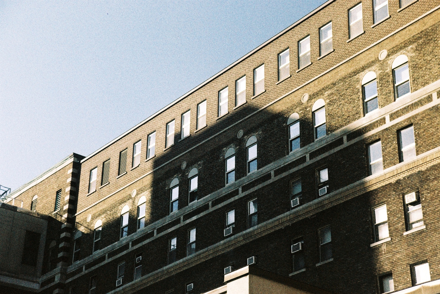 a huge dark brown brick building
that is part of a hospital complex.
it is in the sun,
but the smaller building behind the camera
is casting a big shadow in the middle.