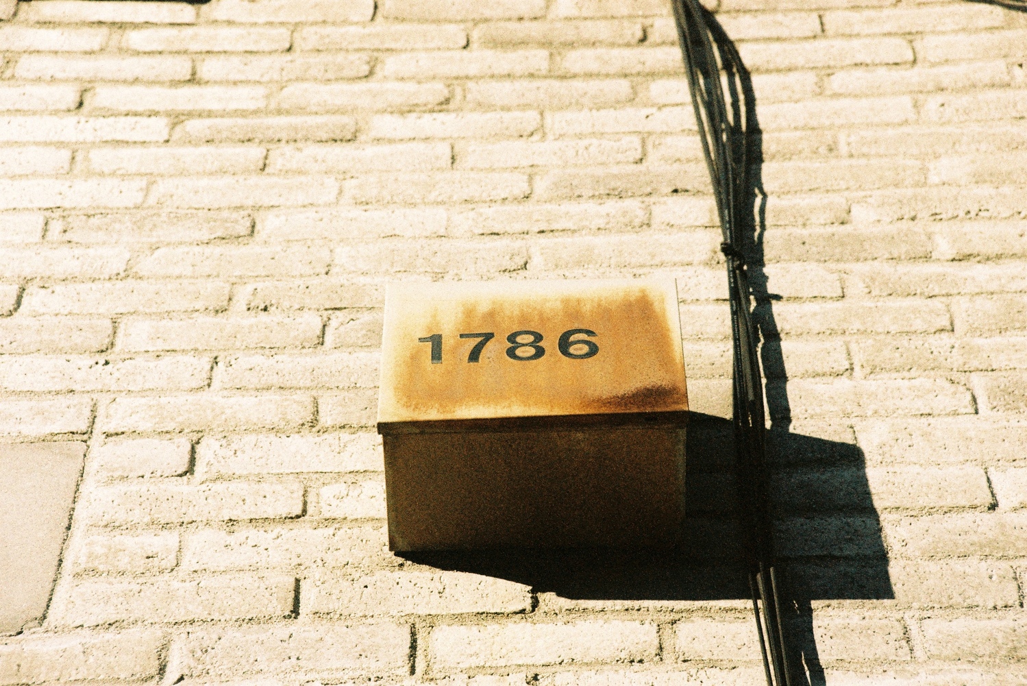 a metal box affixed to a light brick wall
with the number 1786 in black raised lettering on it.
the surface of the metal is rusting a light orange.
just to the right of the box,
a bundle of black wires climbs straight up the wall.