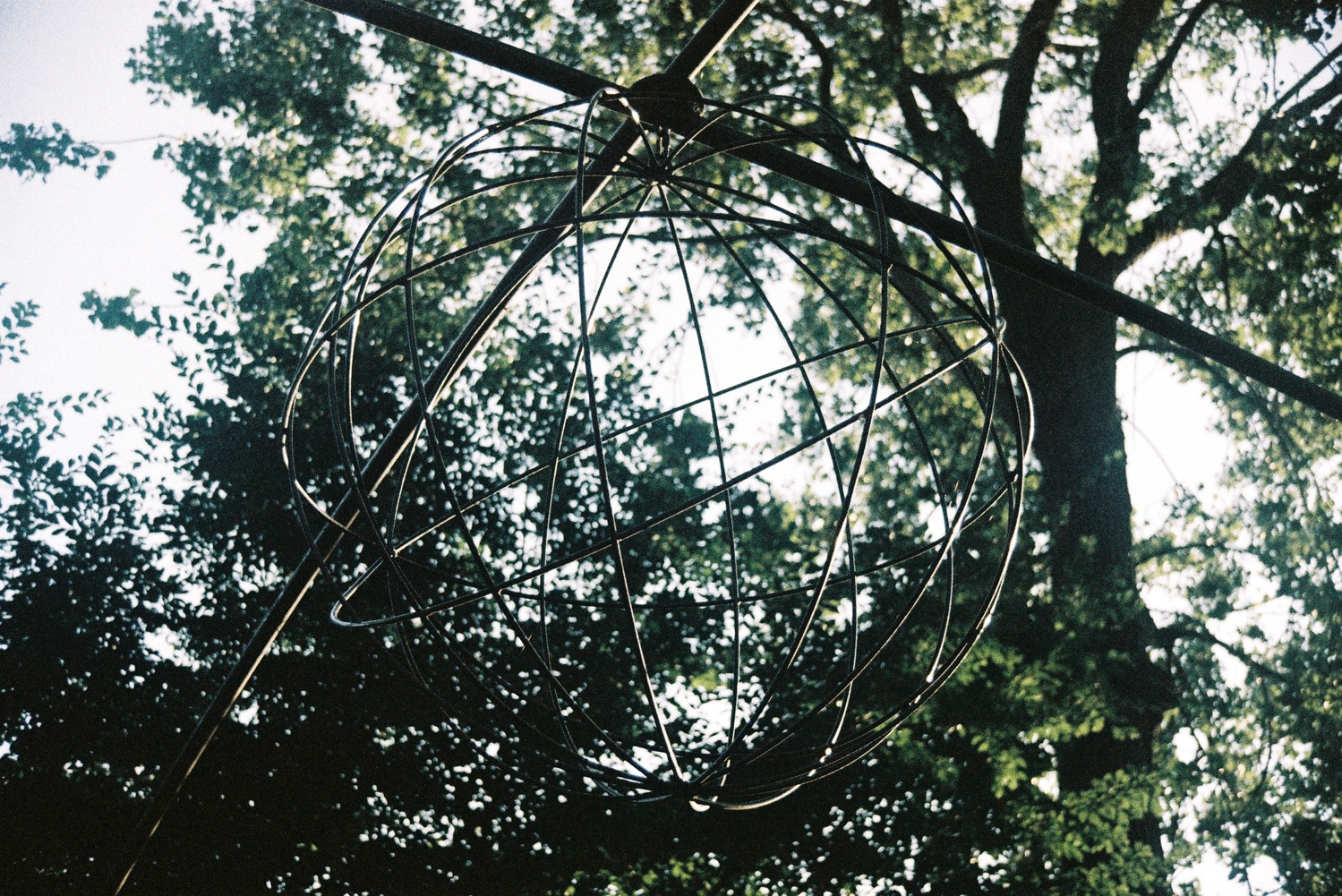 a metal wire sphere
suspended by two crossing metal bars
below some trees.