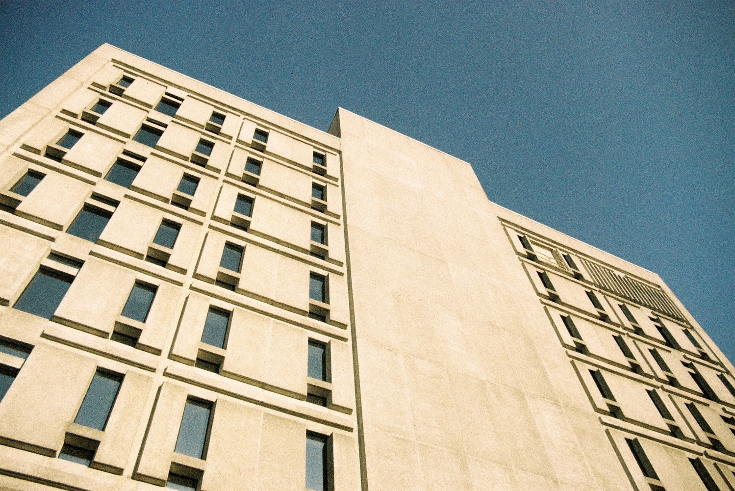 looking up at a concrete building in the sun.
it's got a detailed pattern of depth
around the windows.
the sky is blue.