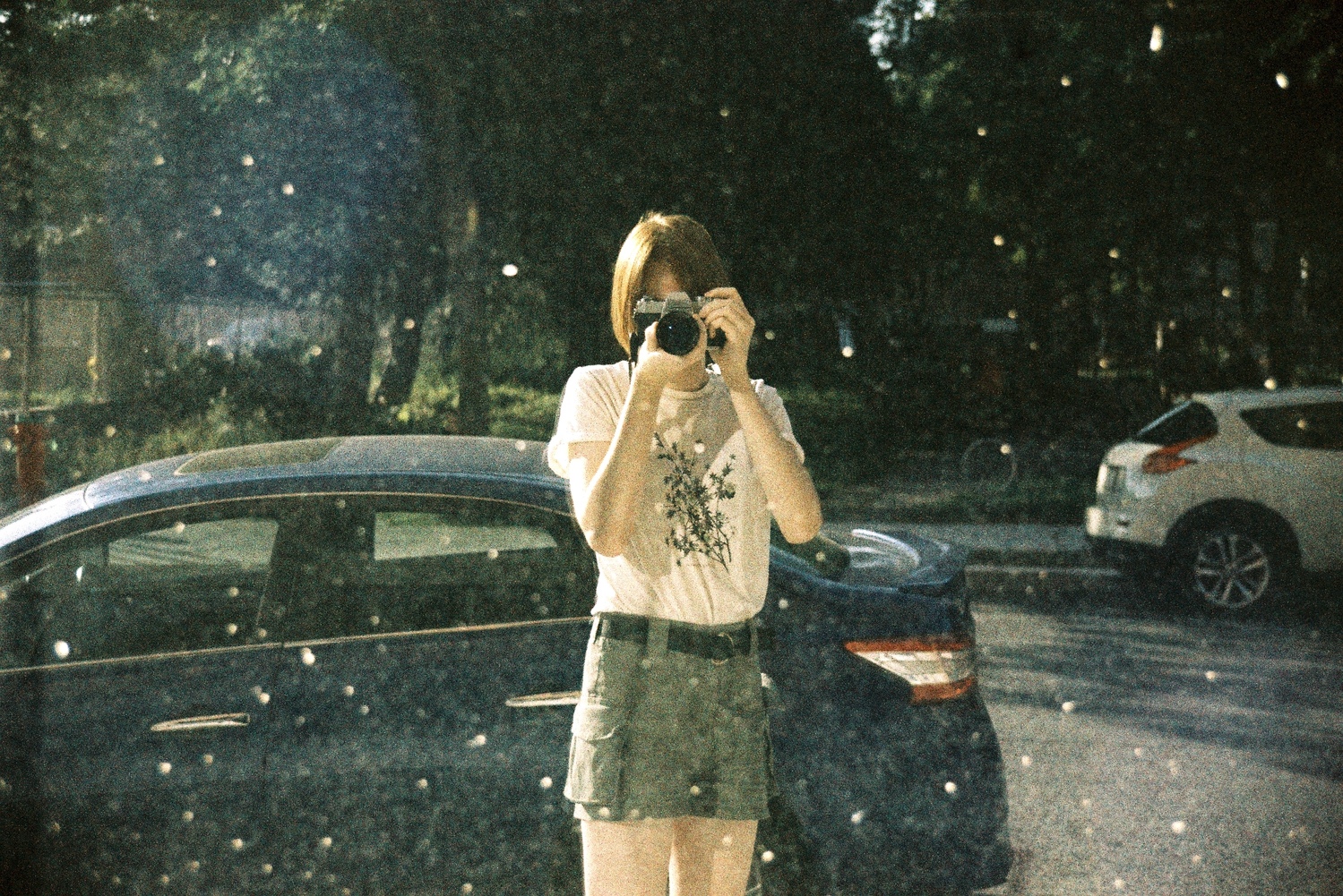 a selfie in some dirty reflective surface I found on the street.
I’m holding up an analog camera to my face
and wearing a light coloured tshirt and green little shorts.
there’s a blue car behind me.