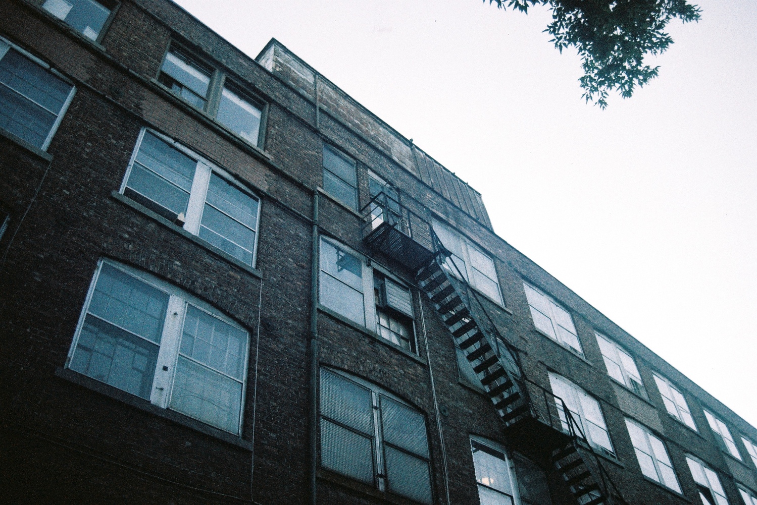 the side of an old brick factory building
with a fire escape going up to one window.
the building is in shadow and the sky is white.