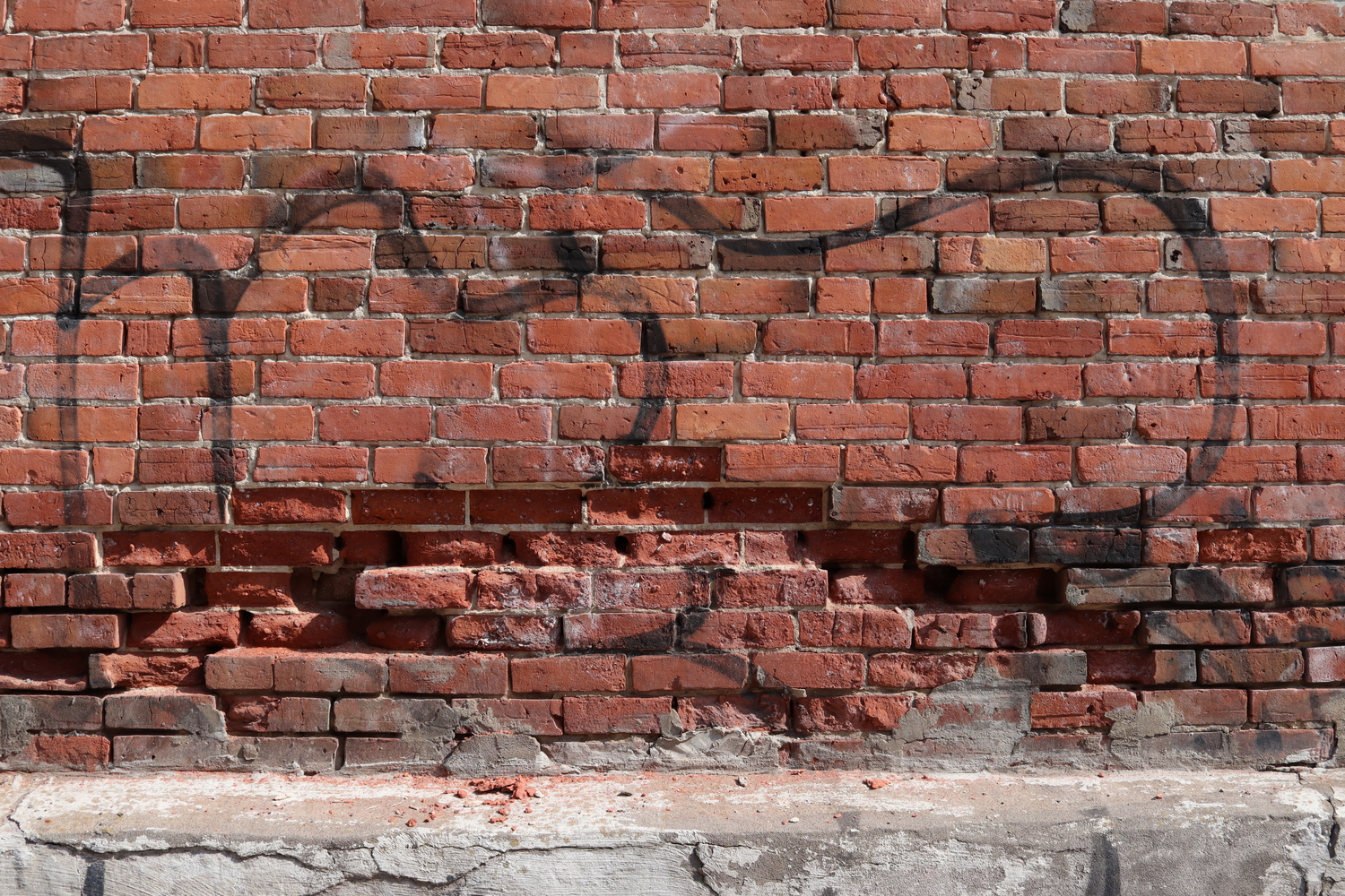 a red brick wall with some faded black graffiti.
in the lower third, some bricks are missing
from the outer layer in an arc shape.
along the bottom is a ledge of conrete
lightly covered in brick dust and chunks
below the missing areas above.