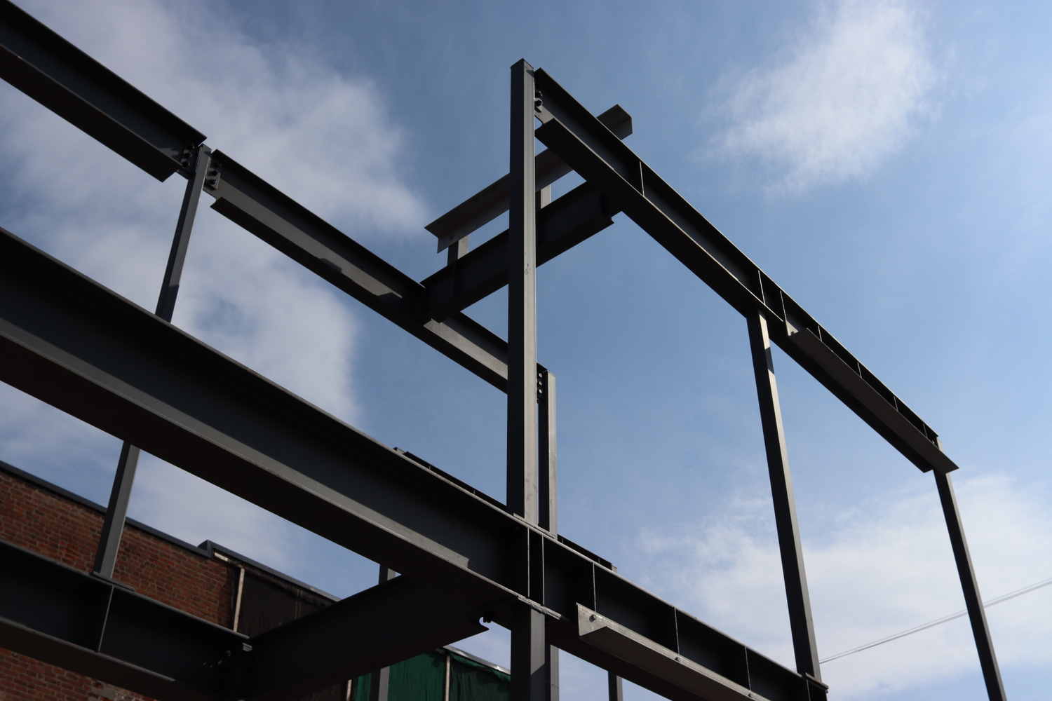 grey steel beams of a building in early construction
on a background of blue sky with some light clouds.
the beams are intersecting at odd points,
implying the final building will not be a simple box.
the sun casts dark shadows into the interiors
of the I-shaped metal.