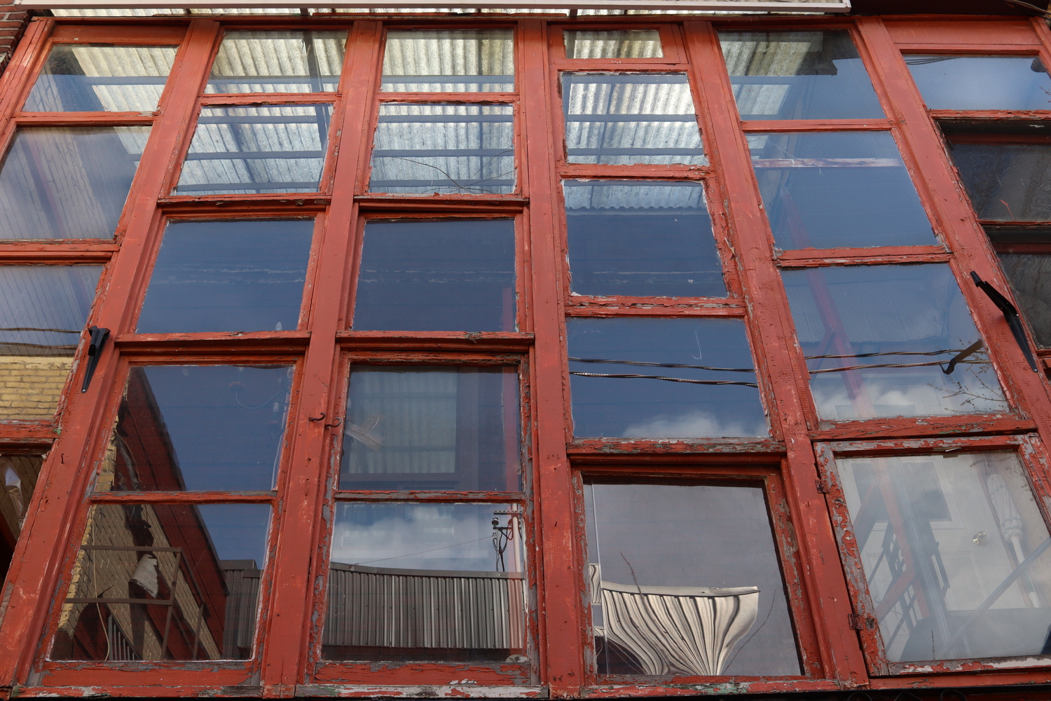 an uneven grid of old wooden-framed windows in an alley.
the red paint on the frames is peeling badly,
completely stripped in some spots.
in the reflections of the lower windows
we see the roofs of the opposite buildings
and hints of clouds in the sky.