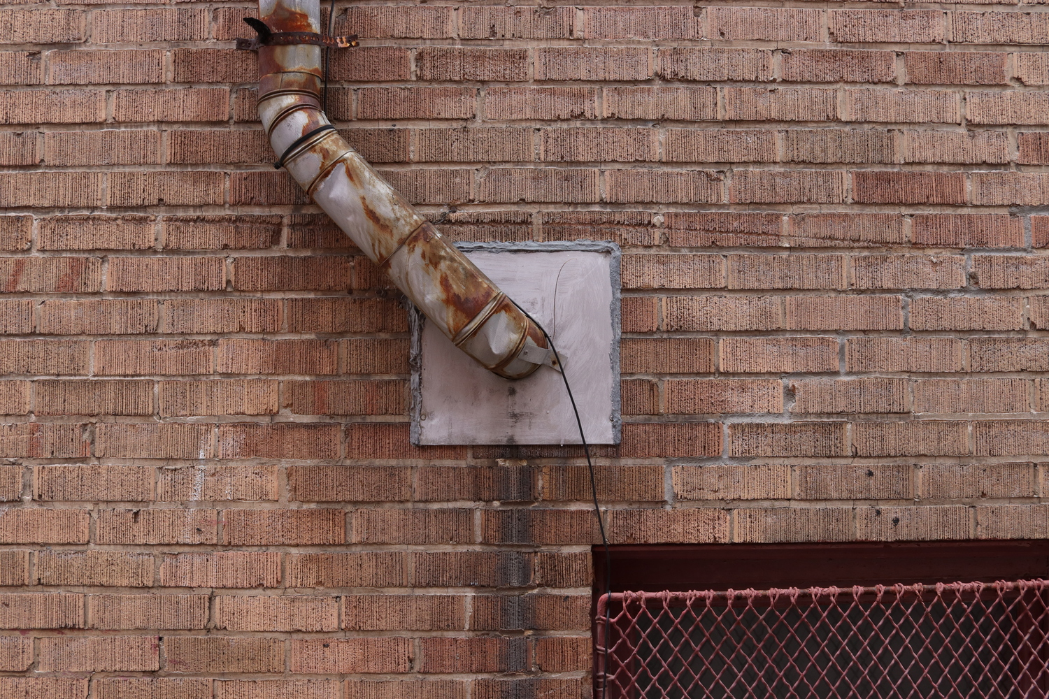 a pipe coming out of a light brown brick wall.
the pipe comes out of a metal square in the centre of the wall,
travels up and left for a bit,
before continuing straight up out of frame.
opposite, in the bottom right,
is the top of a red metal grate in front
of a ground-level window.
the brick below where the pipe enters the wall
is stained dark.