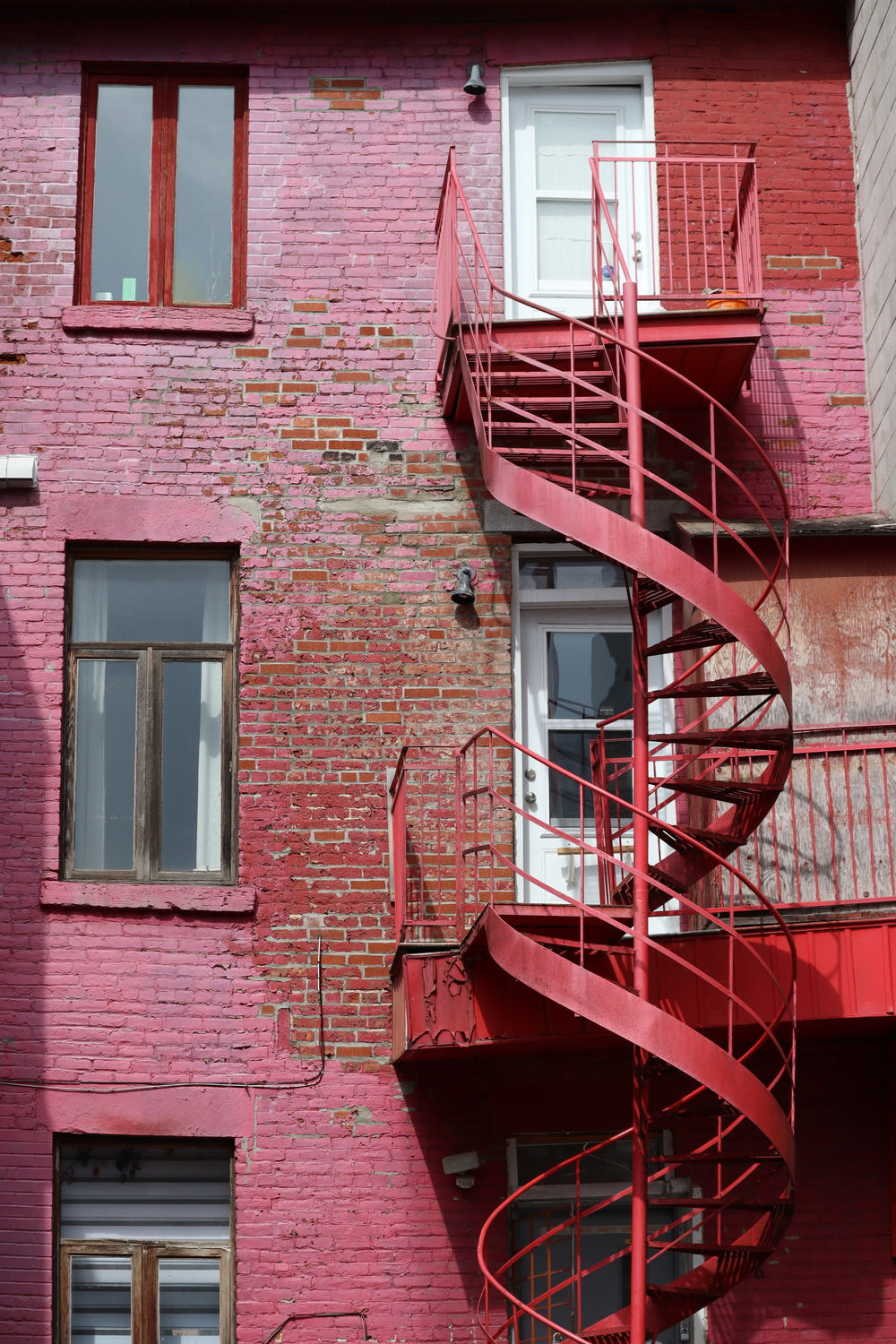 a tall shot of the back of a beautifully coloured building.
the brick wall has been painted a sort of pink,
or at least it's faded to that colour.
there is a splotch in the middle
where the paint has worn off the brick,
along with some stray bricks
elsewhere that have been replaced.
the spiral stairs descending
from the back balconies of two floors
have also been painted red,
but have faded to pink
closer to the top.
everything is a little crooked.
the old wooden-framed windows
on the left,
the more recently replaced doors,
and the balconies.