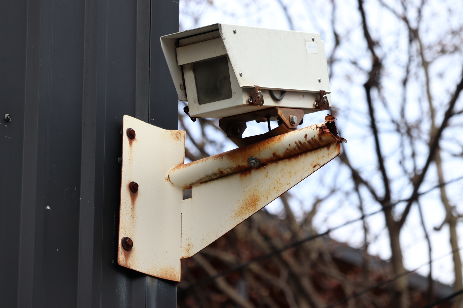 a CCTV camera on the corner
of a black corrugated metal building.
it's mounted on a beige rusting bracket
coming off the wall at a right angle.
it's an old-style boxy camera
with a little hood.
who knows if it's still connected to anything?