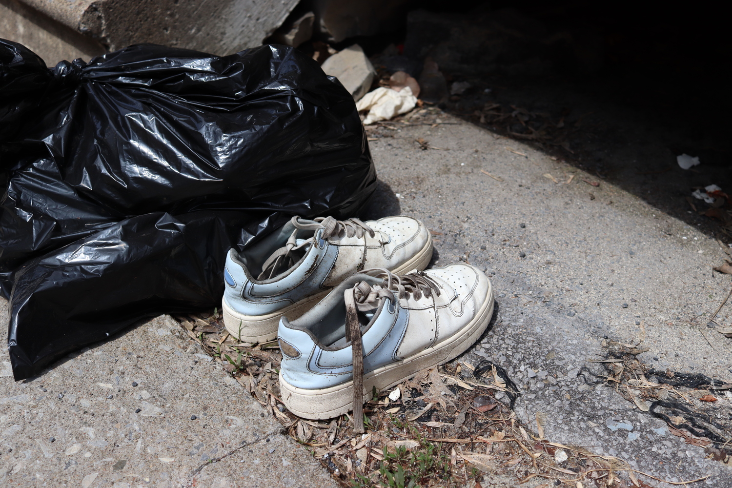 a pair of discarded shoes
on the ground next to a black trash bag.
the shoes are chunky running shoes,
white at the front and baby blue at the back.