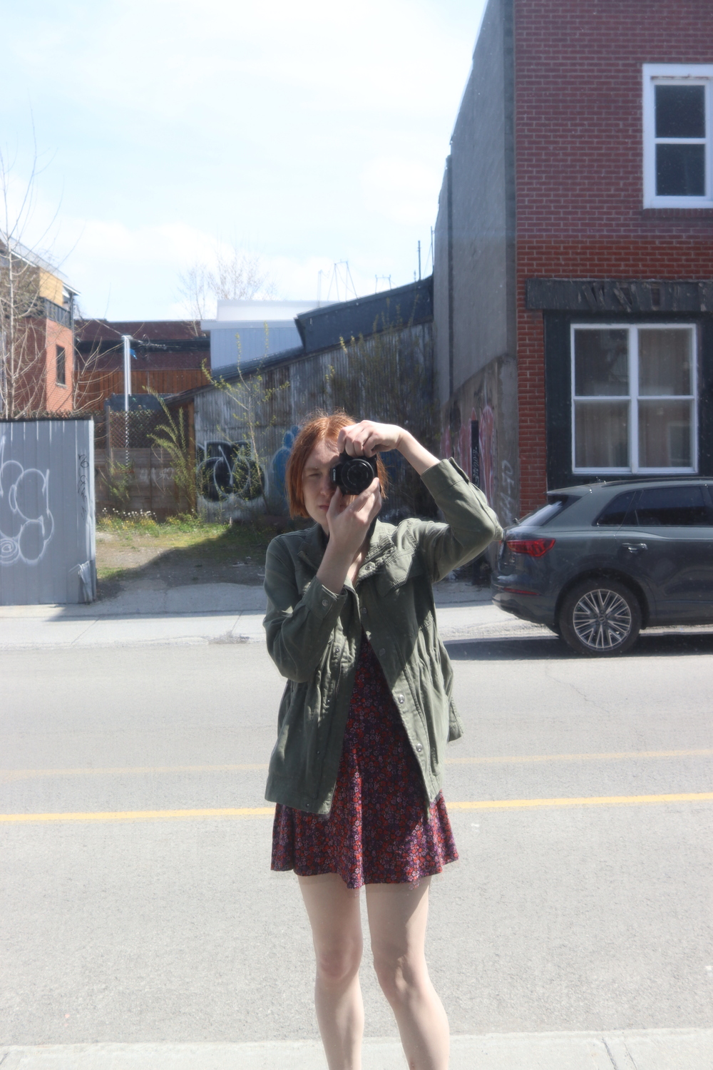 a selfie taken in
a particularly reflective window on the street,
giving everything a slightly offset doubled look
from the layers of glass.
I'm wearing a red and purple floral patterned dress
and a green jacket.
I have red hair
and I'm holding a canon DSLR
up to my face.