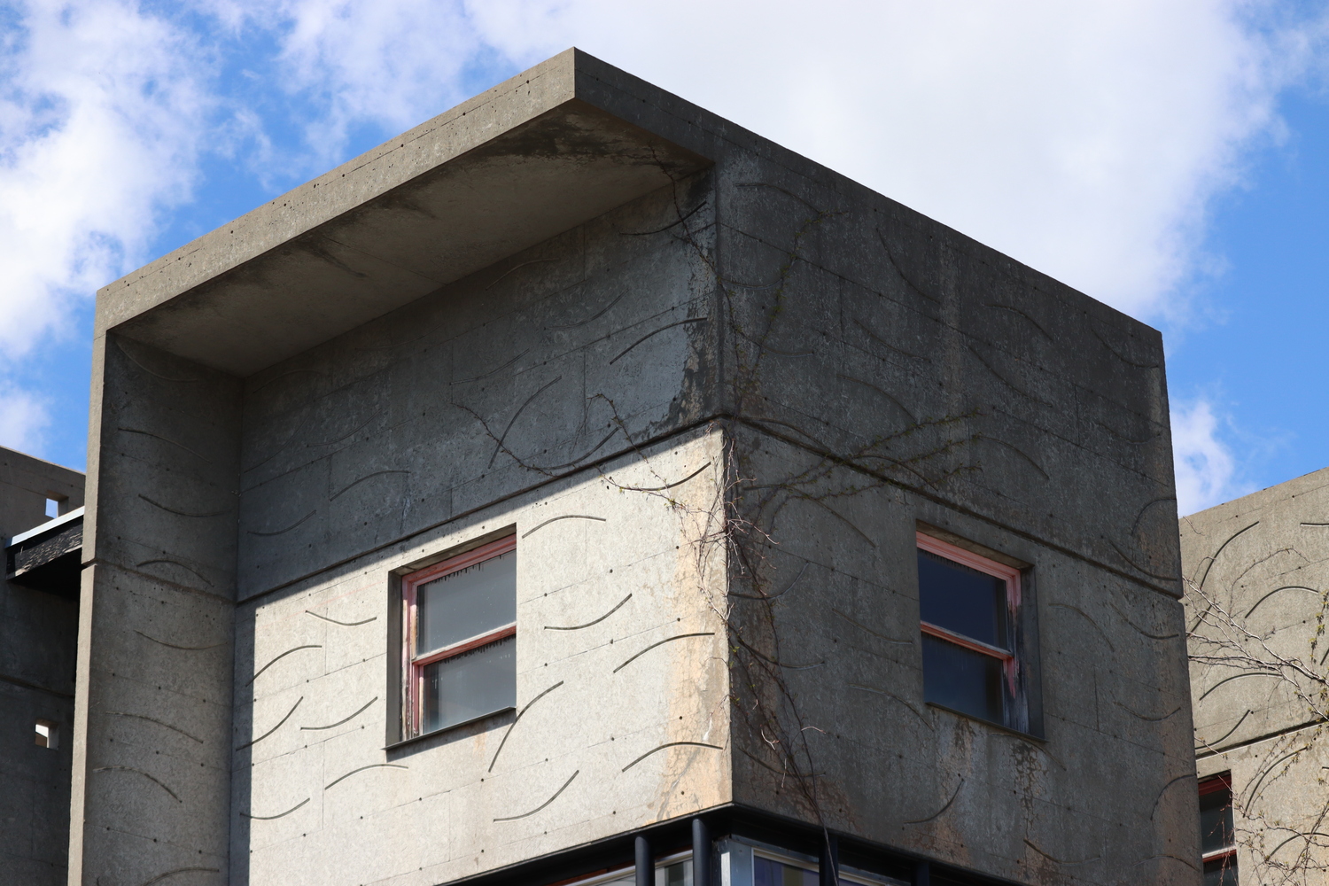 the top cube of a brutalist house,
with one small window
in each the centre of each face.
the left face of the cube
has an overhang above
which continues down the side.
the concrete has large curved grooves
scattered over its surface.
the sun is hitting the left face,
shadowed by the overhang.
behind the cube is a blue sky
with scattered clouds.
