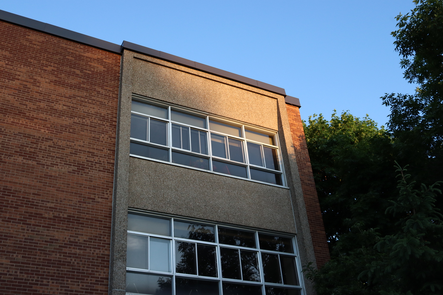 the side of a school building at sunset.
the sun is hitting the top corner.
on the left is the brick wall,
in the centre there are two floors of windows,
and on the right there are green trees.