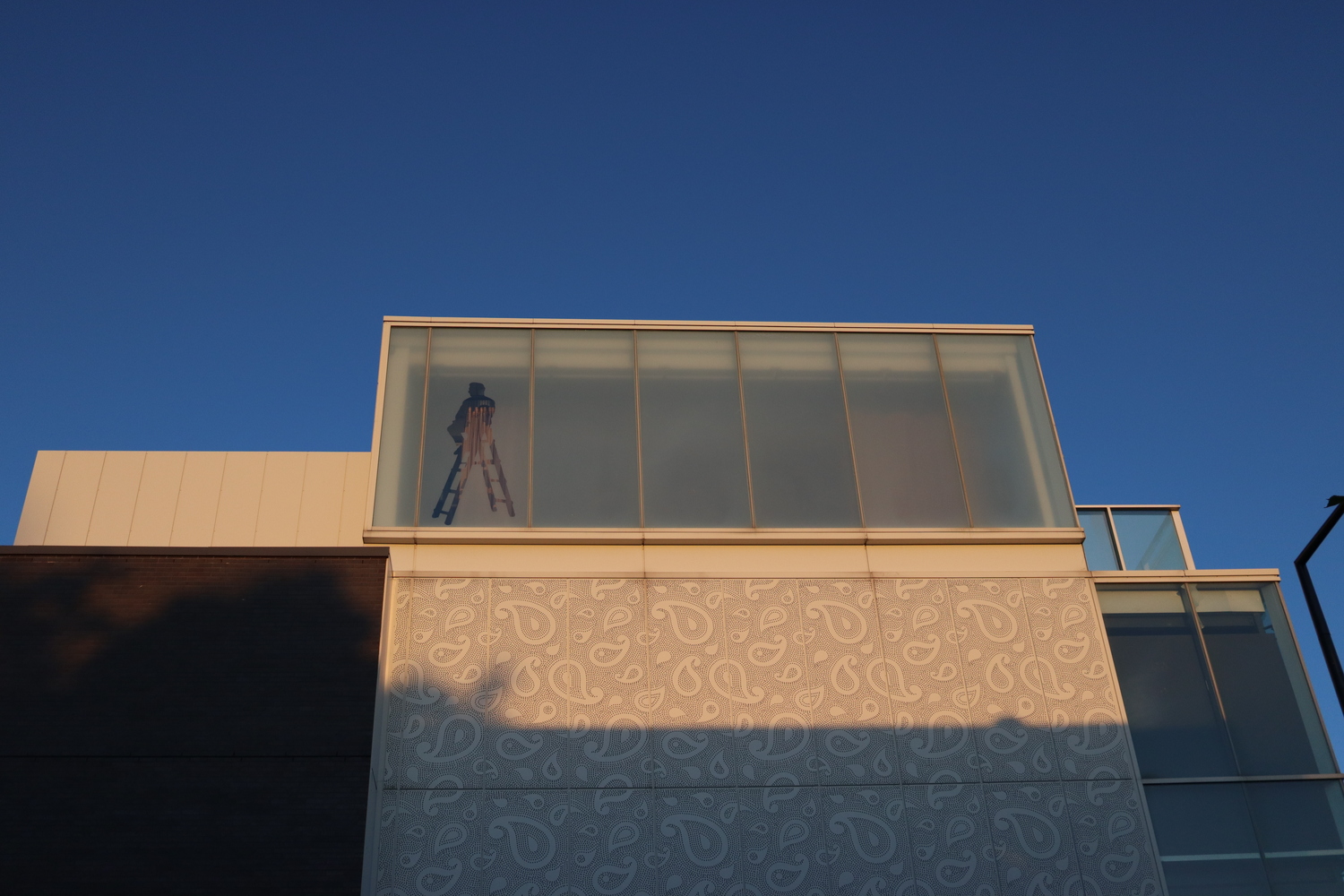 the top of a theatre building at sunset.
the top has a row of frosted glass windows
with an unfrosted silhouette
of a man sitting on a ladder.
below the windows there's a white
swirly teardrop pattern,
which continues down into the shadow of the setting sun.