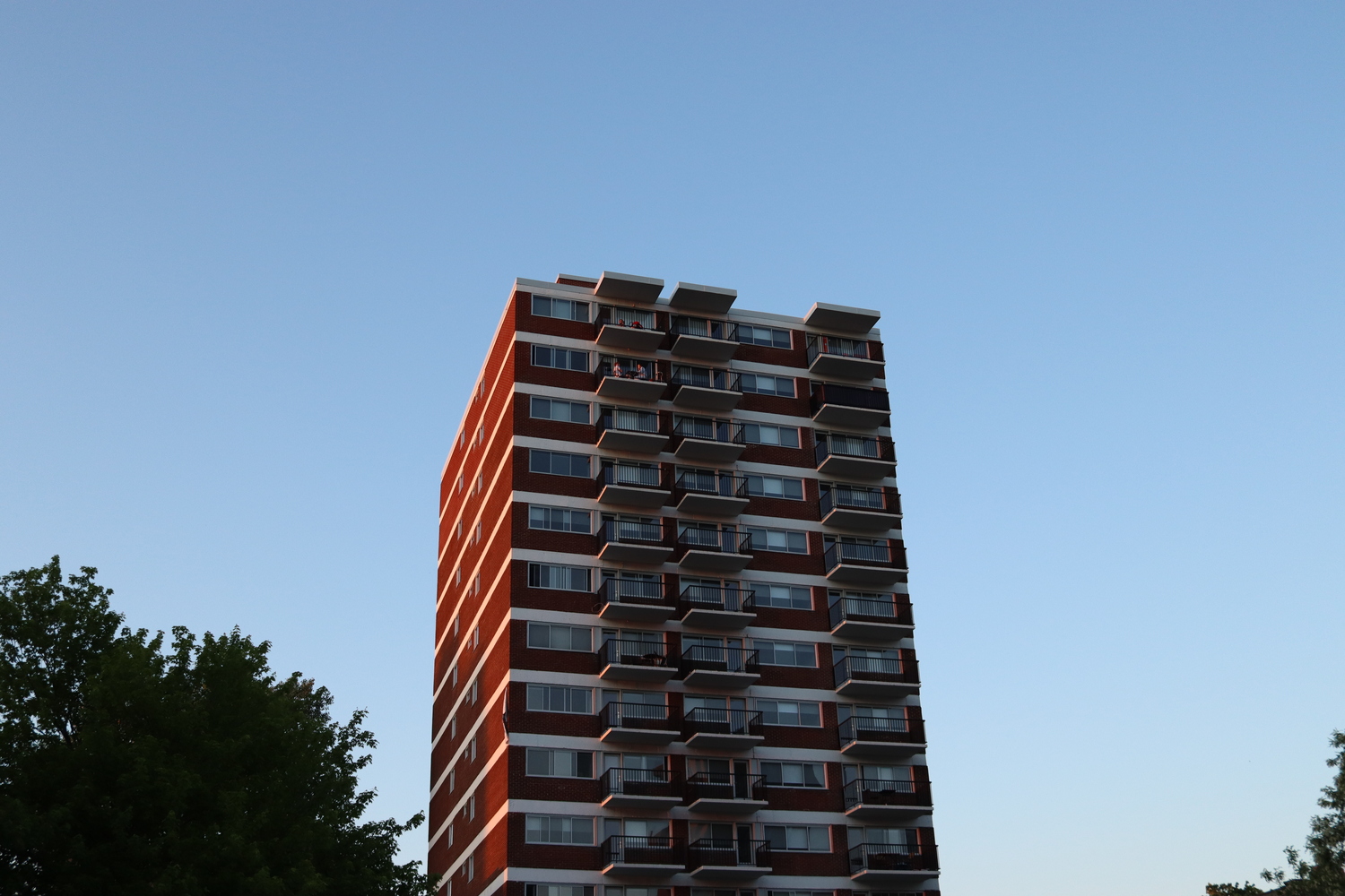 a high-rise apartment building at sunset.
the sun is hitting its left red brick side,
which is broken up by white stripes
between each floor.
the front of the building
has windows and balconies.
some of the balconies near the top
have people out on them.