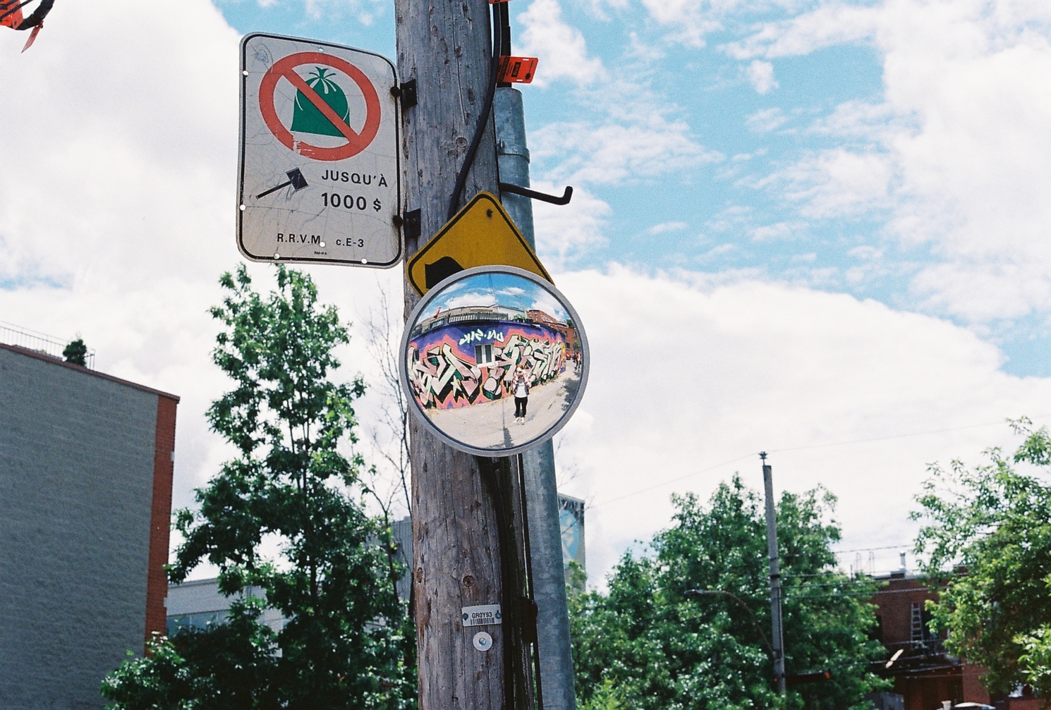 a selfie in a convex mirror
mounted on a telephone pole.
I appear quite small in the mirror.
I'm standing in front
of a building with
colourful graffiti on it.
behind the mirror
is a yellow diamond road sign,
and on the side of the pole
is a no trash sign
with a fine of up to $1000.
behind is a blue cloudy sky.