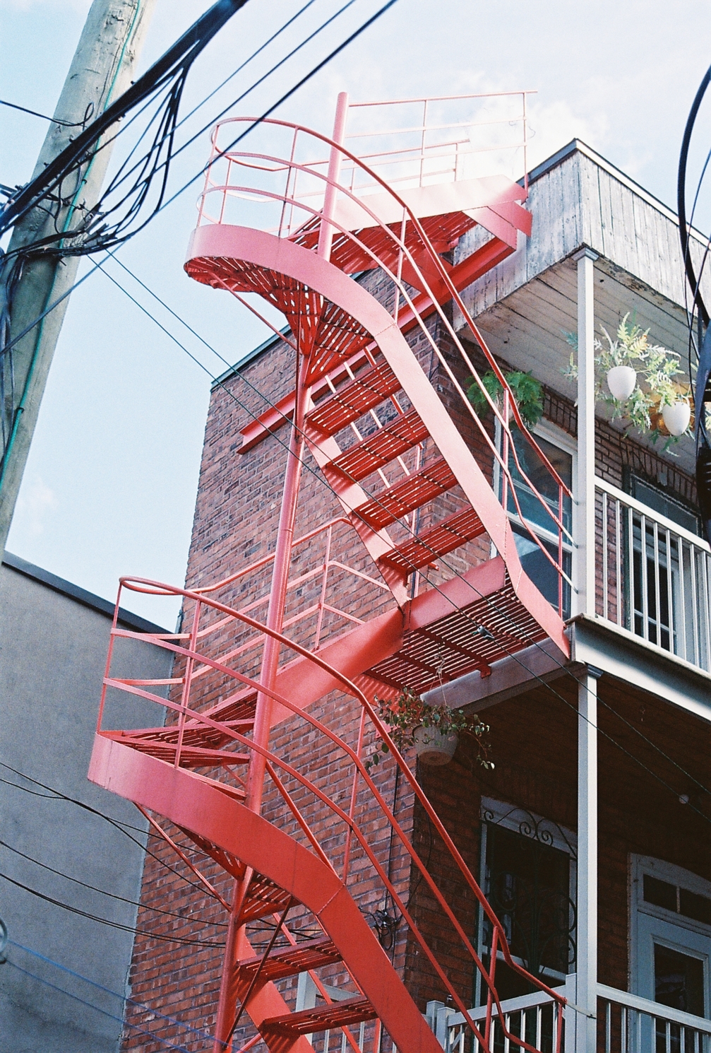 a painted red metal fire escape spiral staircase
climbing up the side of a narrow brick building.
it goes all the way to the roof,
where it becomes over-exposed
and starts to blend into the sky behind it.