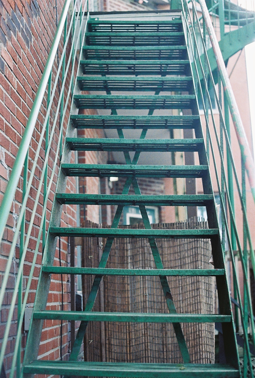 a green metal staircase
viewed as if about to climb it,
hugging the back of a brick building.